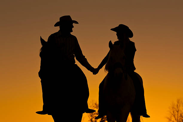 Couple horseback riding side by side, silhouetted against the setting sun.  Cowboy hats, orange background.
